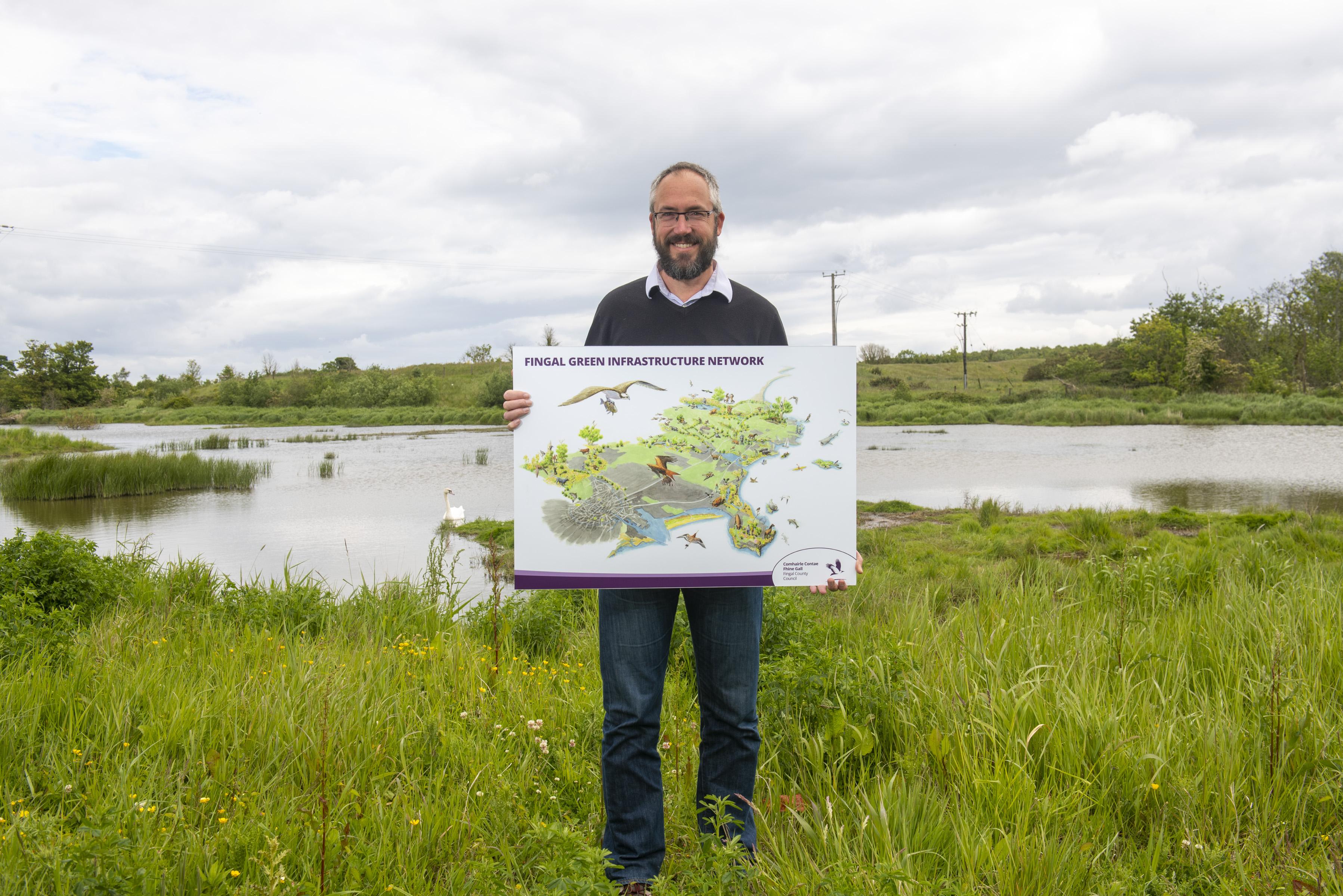 bearded man holding illustration of fingal county stands next to water with swans in background