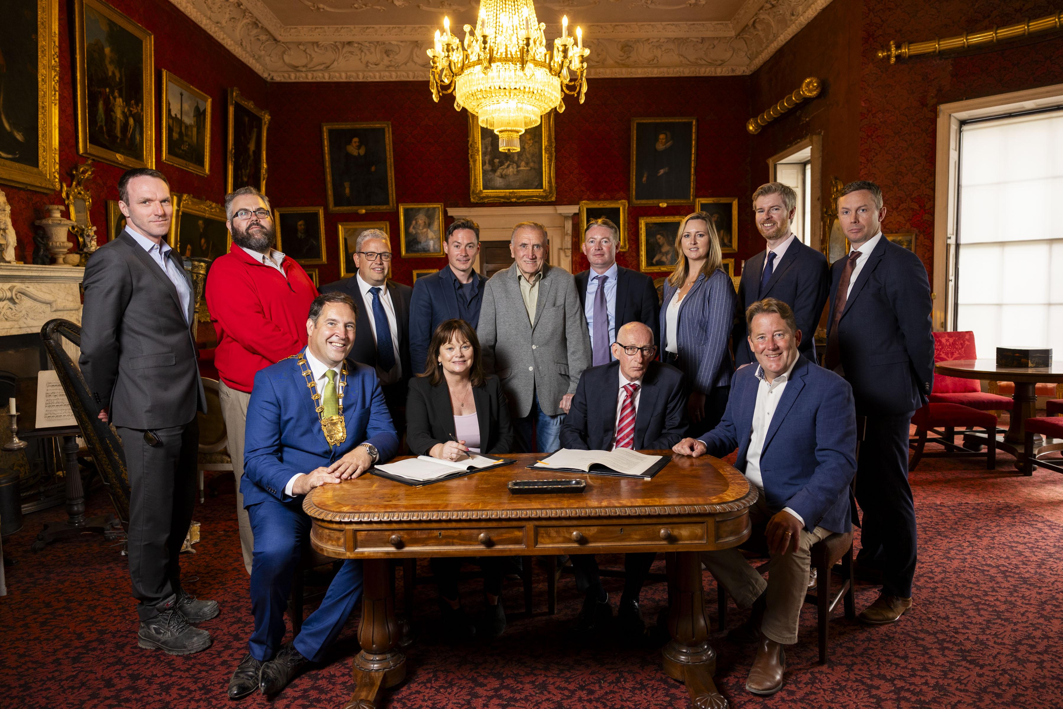 large group of men and women standing around conference table as contract signing happens